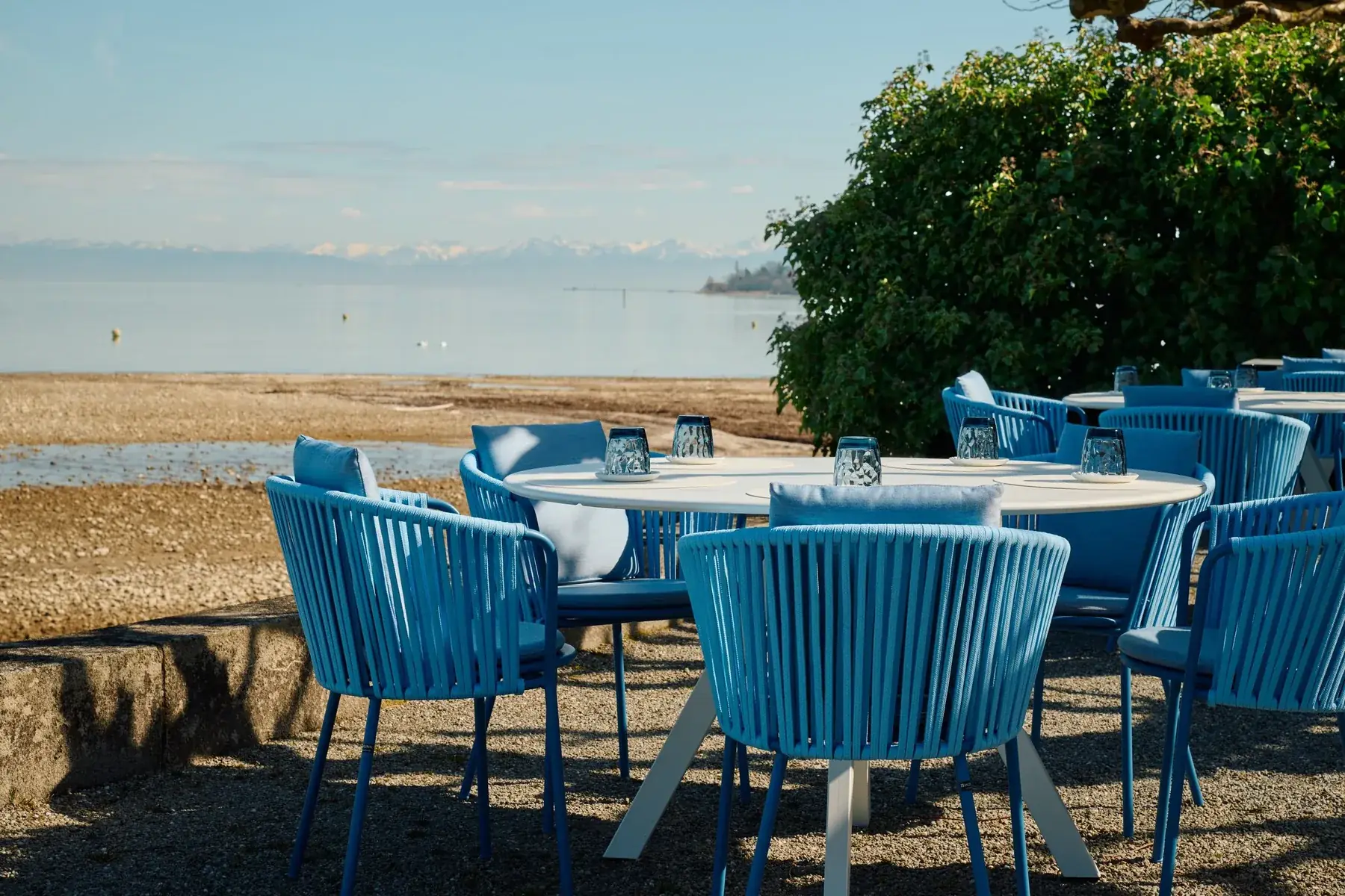 Blaue Stühle auf der Terrasse am Mittag, dahinter der Bodensee und die Alpen