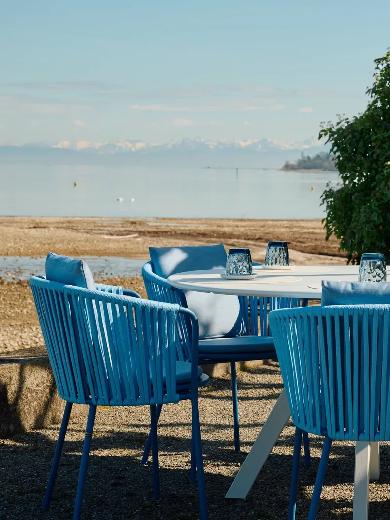 Leere blaue Stühle am Wasser, dahinter die Alpen